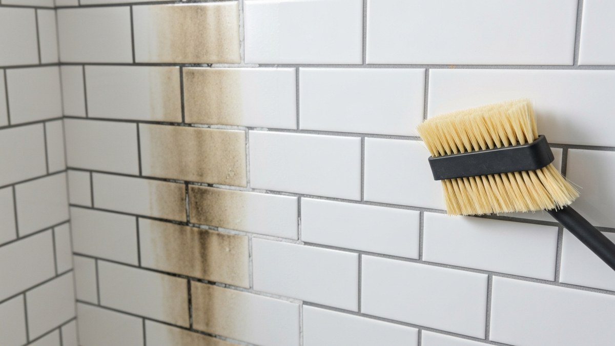 How to clean grout stains: discolored grout lines on white wall tile beside a scrub brush