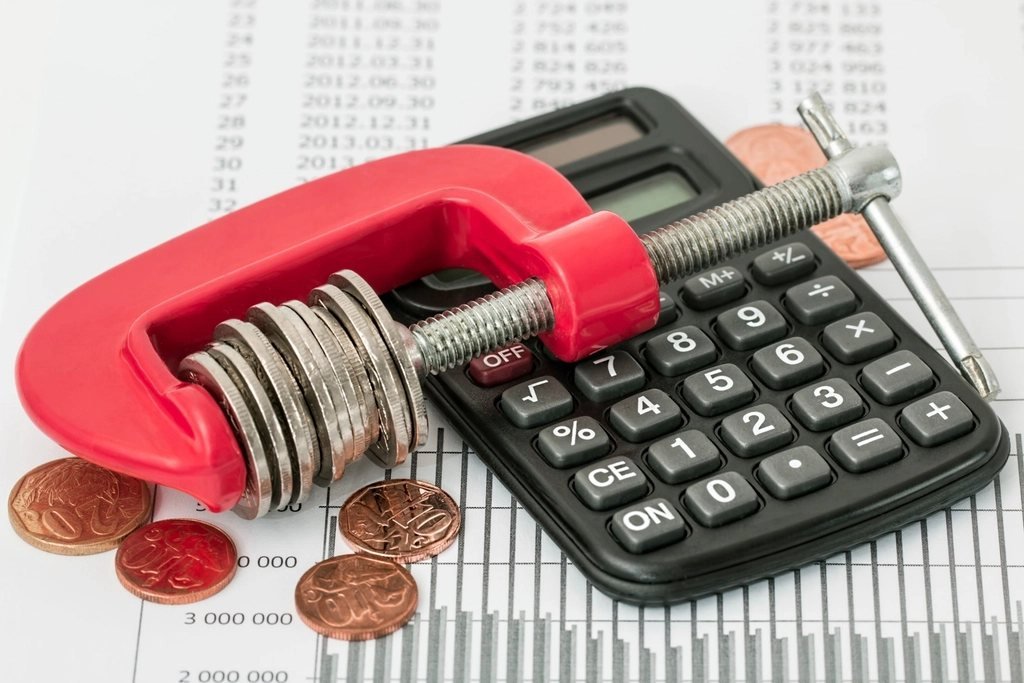 Coins squeezed in a clamp beside a calculator, illustrating how lowball bids squeeze budgets