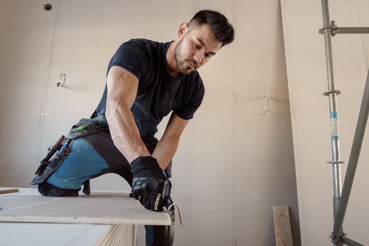 A worker scoring and cutting a sheet of drywall on a jobsite with hung drywall behind